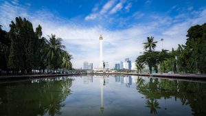 National Monument (Monas) in Jakarta, Indonesia, reflecting in water with blue skies and cityscape.