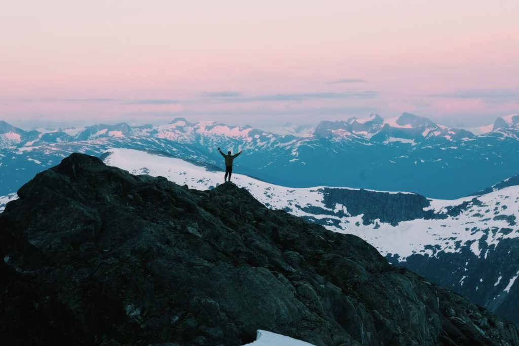 Silhouette of a person with arms spread on a snowy mountain top during a pink sunset.