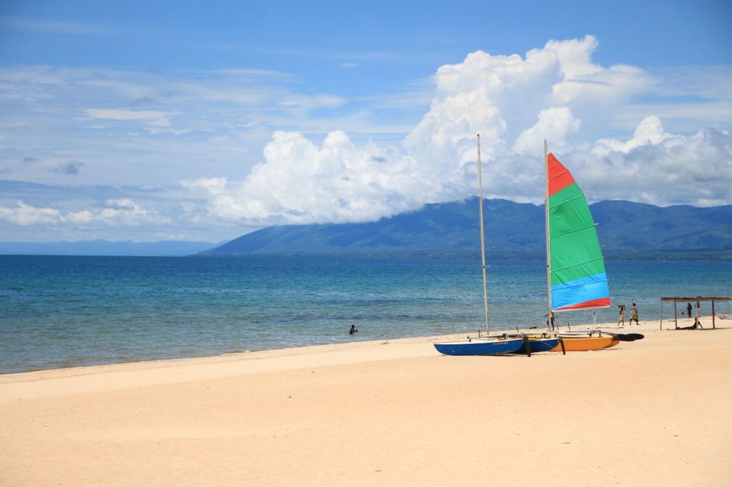 Vibrant sailboats rest on a sunny Lake Malawi beach with mountains in the background.