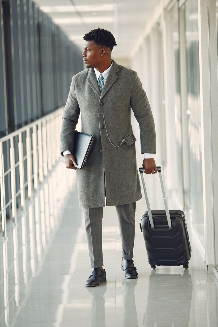 Full length of stylish serious businessman in elegant gray suit walking with suitcase and suitcase in modern airport hallway while looking away pensively